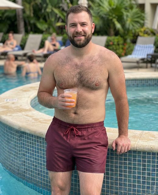 Man wearing burgundy swim shorts holding a drink at a resort pool